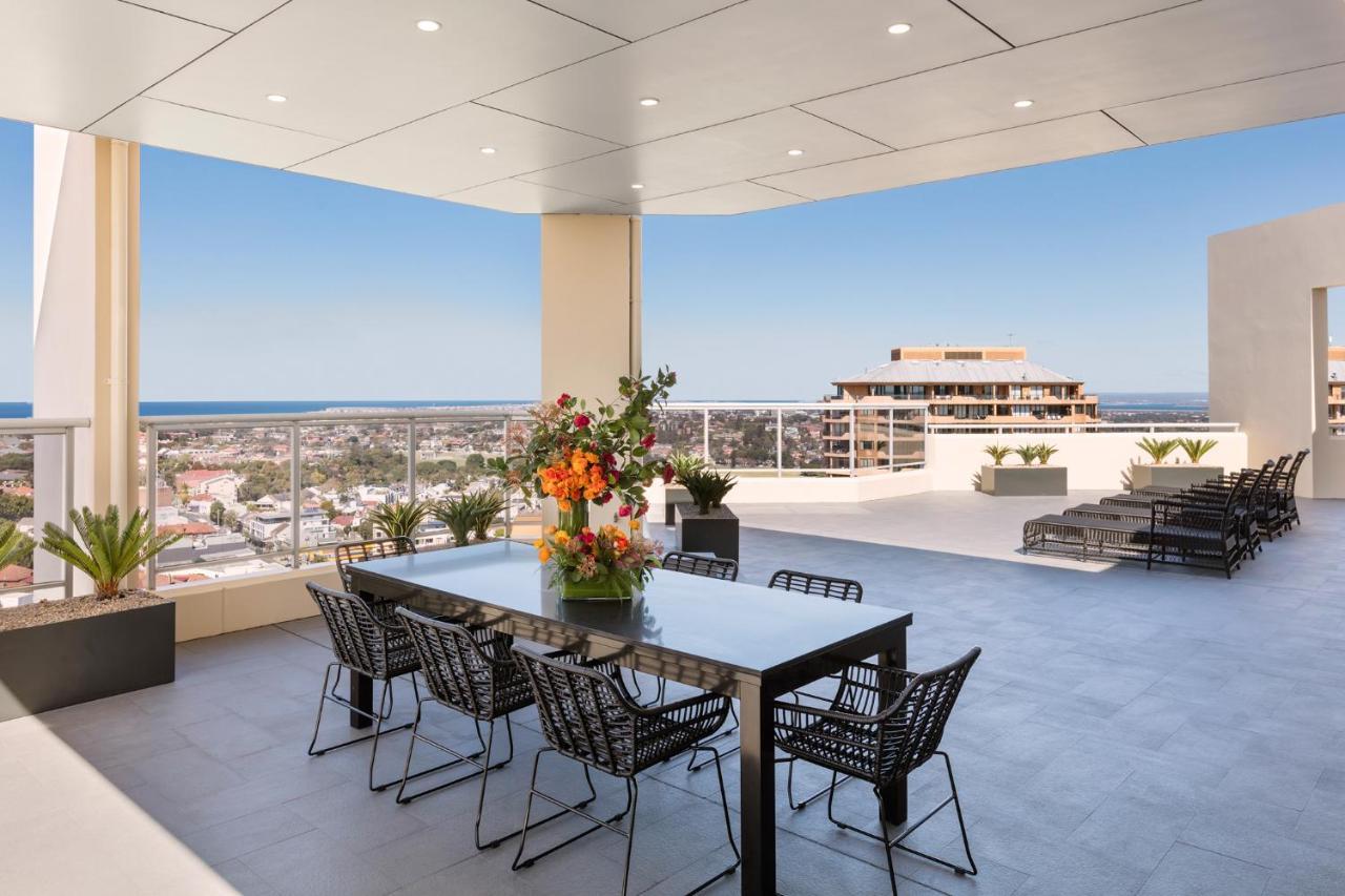 Meriton Suites Bondi Junction Sidney Dış mekan fotoğraf The photo shows a stylish outdoor terrace or balcony area, featuring a long dining table surrounded by several black wire chairs. The table has a centerpiece of vibrant flowers. The space is well-lit, with a clear view of the horizon in the backgroun