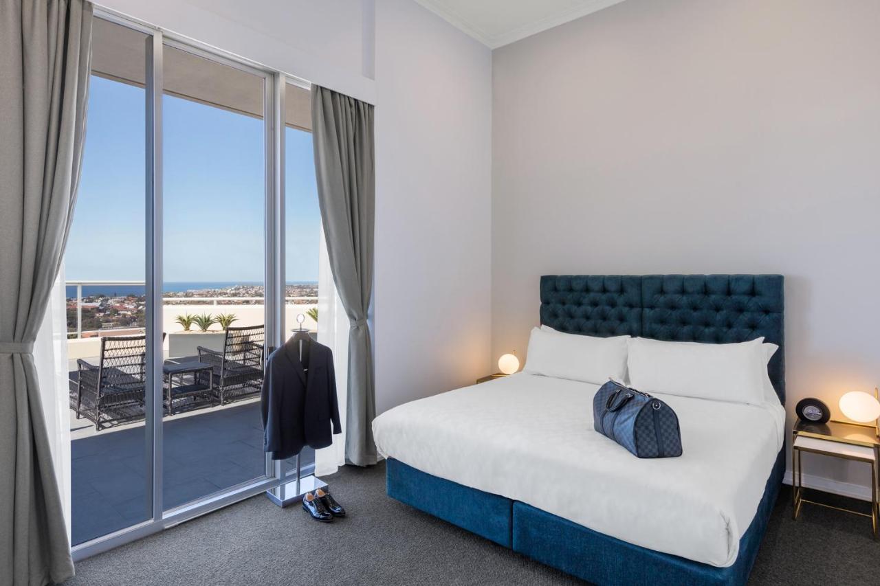 Meriton Suites Bondi Junction Sidney Dış mekan fotoğraf The photo shows a stylish bedroom featuring a large bed with a tufted blue headboard. There's a neatly organized outfit consisting of a suit hanging on a stand, along with shoes placed on the floor. The room has large windows that provide a view of t