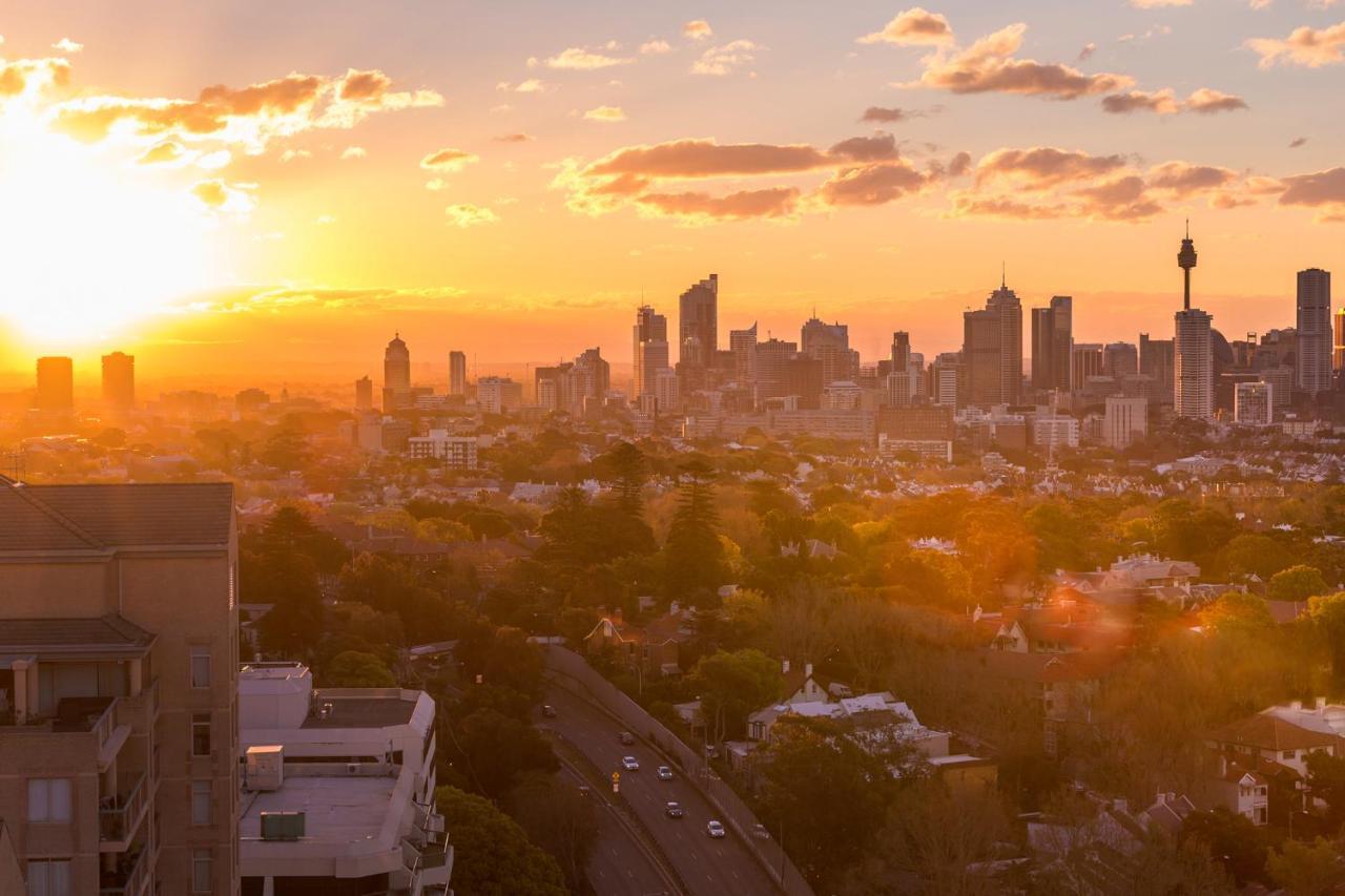 Meriton Suites Bondi Junction Sidney Dış mekan fotoğraf The photo shows a panoramic view of a city skyline at sunset. The sun is low on the horizon, casting a warm golden light across the buildings. Tall skyscrapers are silhouetted against the colorful sky, which features hues of orange, pink, and yellow,
