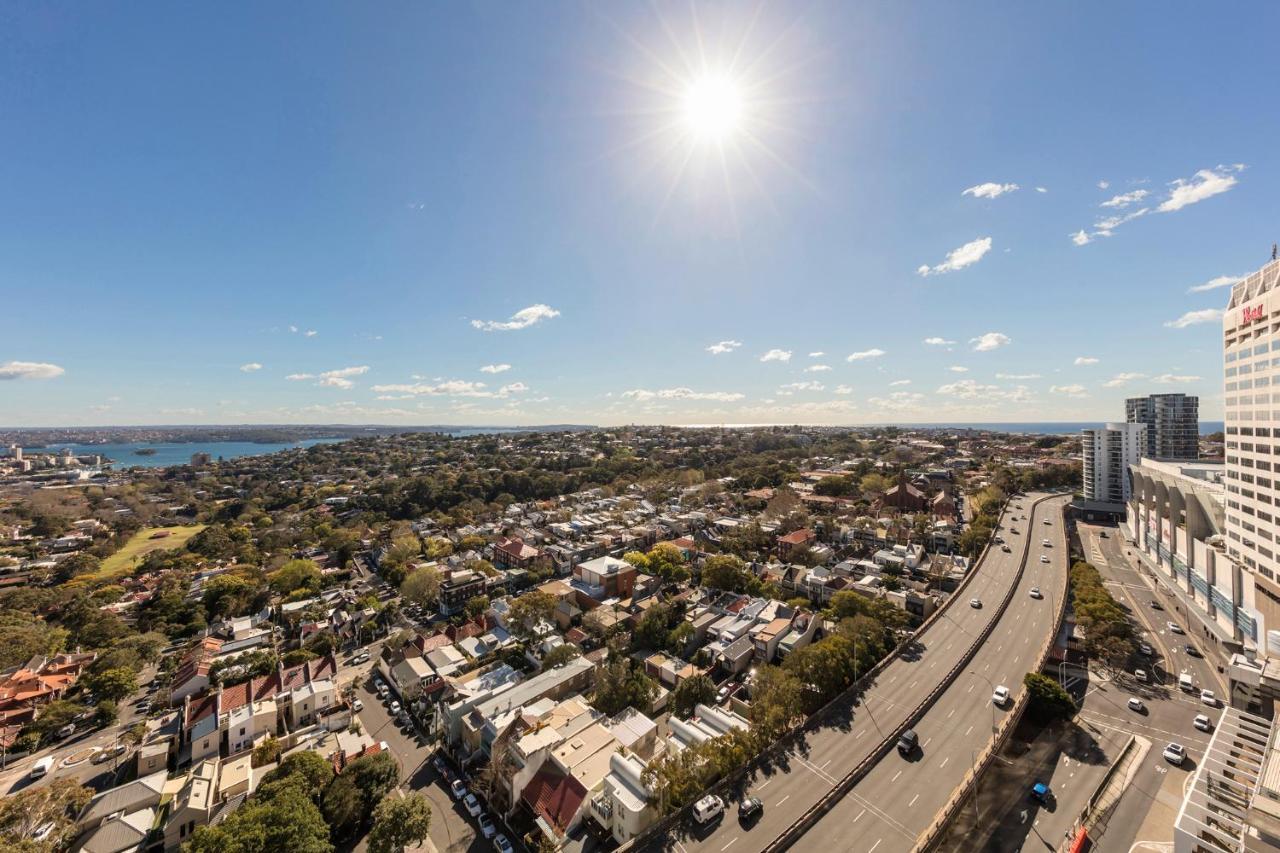 Meriton Suites Bondi Junction Sidney Dış mekan fotoğraf The photo shows a panoramic view of a city landscape under a clear blue sky. In the foreground, there is a road with vehicles traveling along it, bordered by trees and buildings. In the background, you can see a residential area with various houses, 