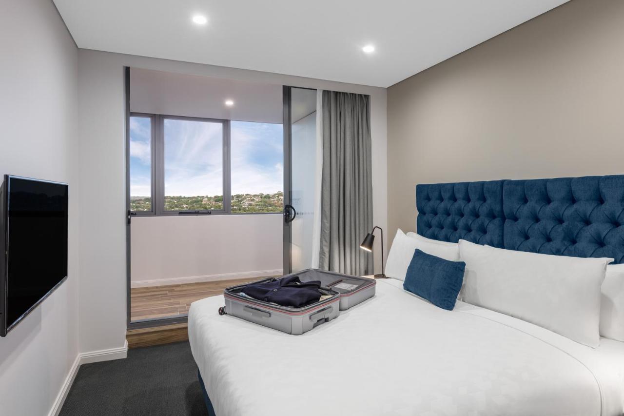 Meriton Suites Bondi Junction Sidney Dış mekan fotoğraf The photo shows a modern hotel room featuring a double bed with a tufted blue headboard. There is a neatly arranged suitcase placed open on the bed, indicating that someone may be unpacking. The room has light-colored walls and a window allowing natu