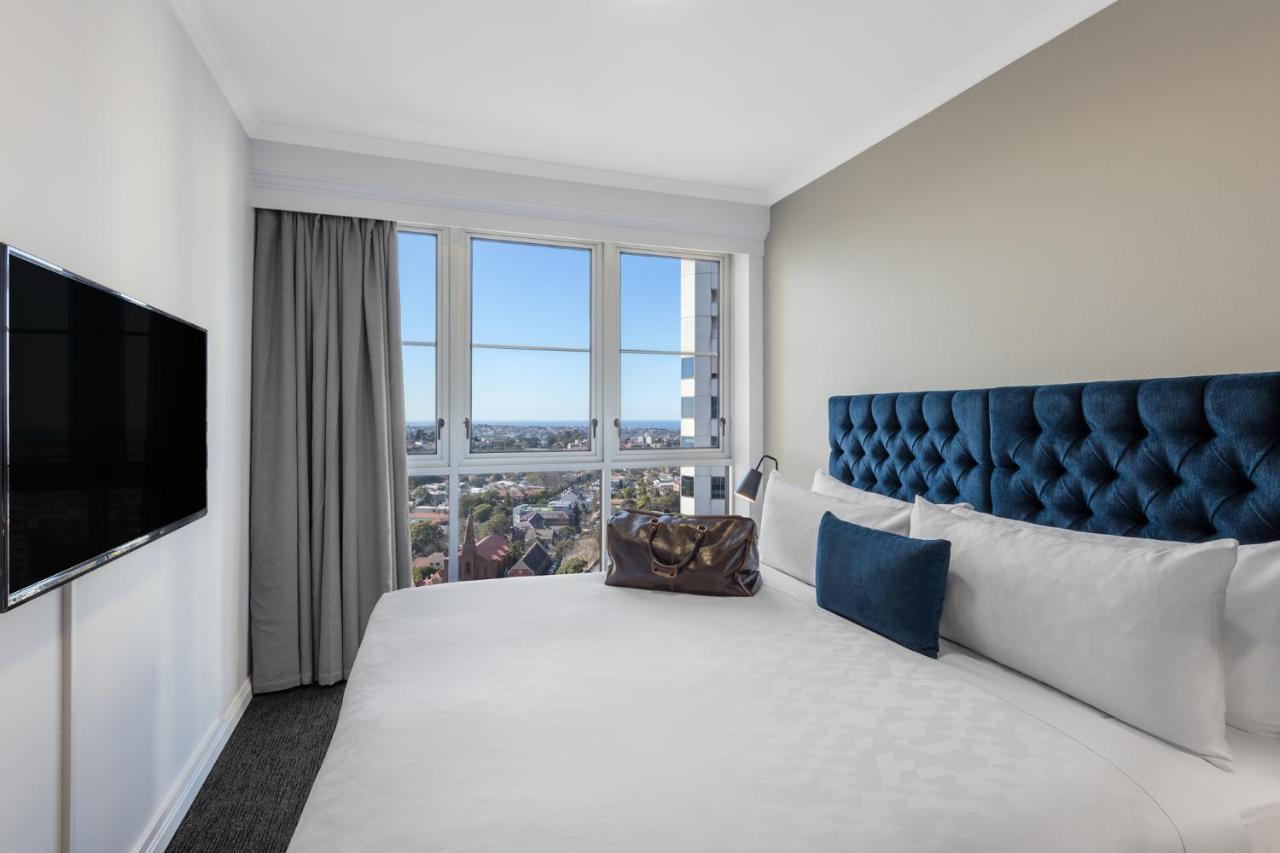 Meriton Suites Bondi Junction Sidney Dış mekan fotoğraf The photo shows a modern hotel room featuring a large bed with a tufted blue headboard. Beside the bed, there is a stylish bag placed on the bed. The room has large windows that provide a view of the outside, allowing natural light to fill the space.