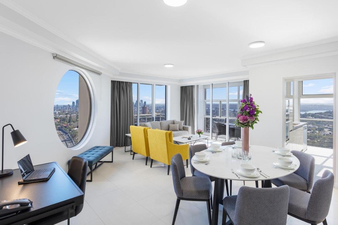 Meriton Suites Bondi Junction Sidney Dış mekan fotoğraf The photo shows a modern and bright interior space, likely a living room or dining area. It features large windows that offer a panoramic view of the city skyline. The furnishings include a round dining table set for several people, with plates and g
