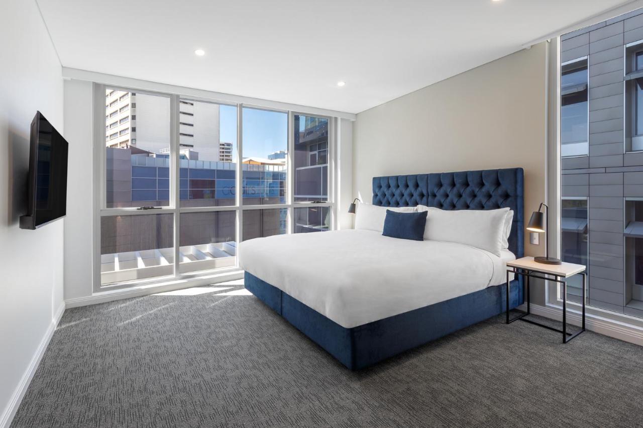 Meriton Suites Bondi Junction Sidney Dış mekan fotoğraf The image shows a modern hotel room with a large, comfortable bed featuring a blue upholstered headboard. The room has large windows that allow natural light to enter, providing a good view of the surrounding buildings. There is a small bedside table