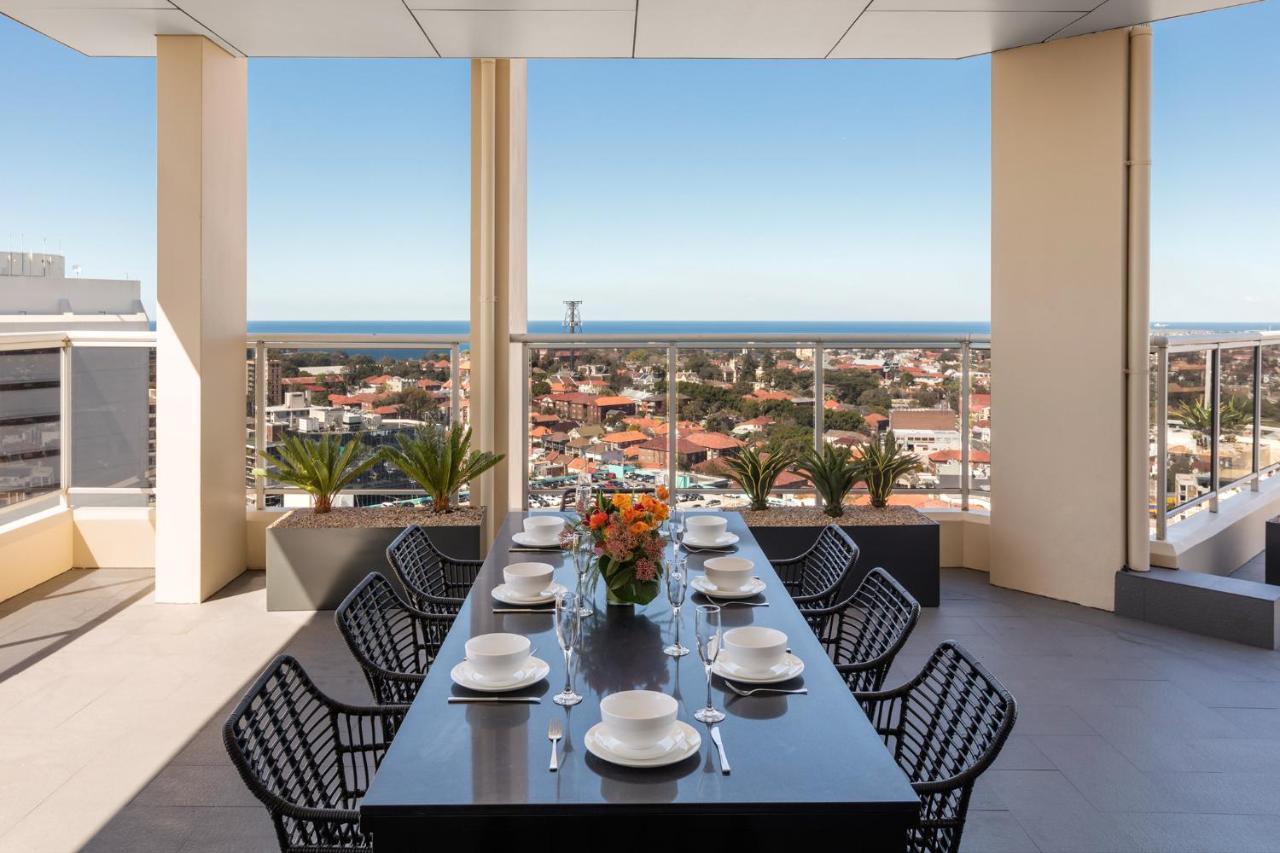 Meriton Suites Bondi Junction Sidney Dış mekan fotoğraf The photo shows a modern outdoor dining area situated on a balcony or terrace with a stunning view. There is a long rectangular table set with plates, glasses, and cutlery, ready for a meal. The table is surrounded by sleek, black wicker chairs. In t