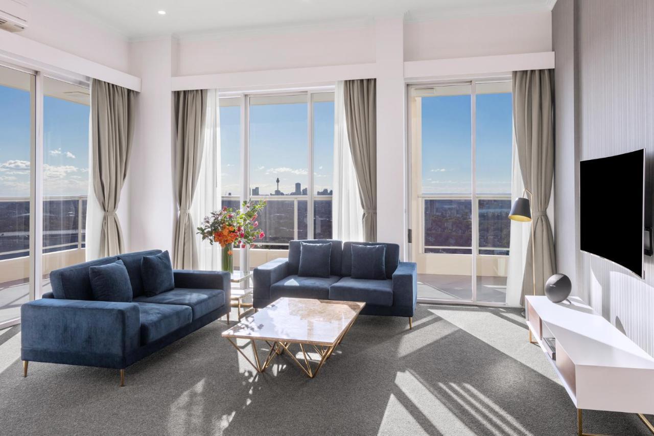 Meriton Suites Bondi Junction Sidney Dış mekan fotoğraf The photo shows a modern living room with large windows that allow natural light to fill the space. There are two blue sofas arranged around a coffee table with a glass top and gold legs. A floral arrangement adds a touch of color, placed on a nearby