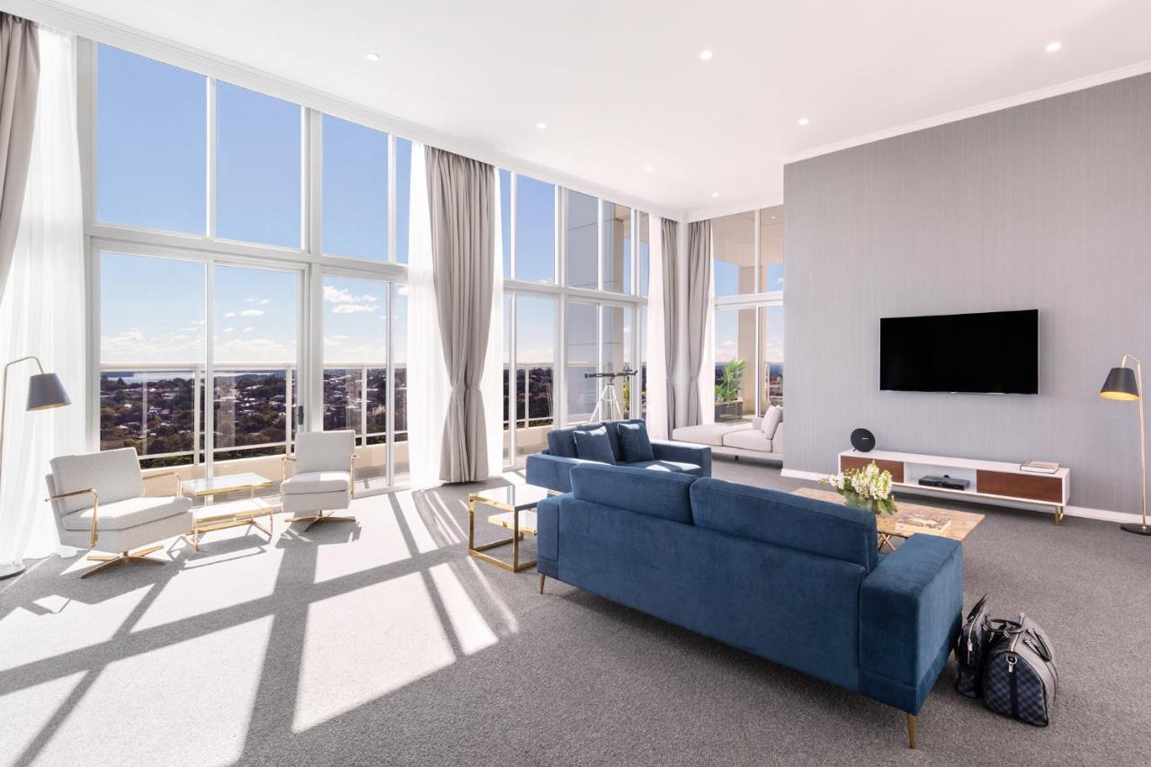 Meriton Suites Bondi Junction Sidney Dış mekan fotoğraf The photo shows a bright and modern living room with large floor-to-ceiling windows that provide a view of the outdoors. The space features a blue sofa in the center, complemented by light-colored armchairs nearby. There are light curtains hanging by
