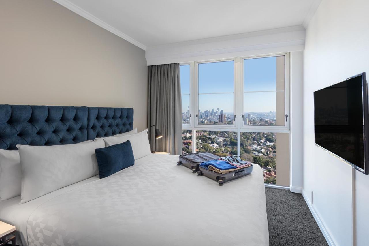 Meriton Suites Bondi Junction Sidney Dış mekan fotoğraf The photo shows a modern hotel bedroom featuring a king-sized bed with a blue tufted headboard. The bed is neatly made with white linens and a decorative blue pillow. There is a large window that offers a view of the city skyline, allowing natural li