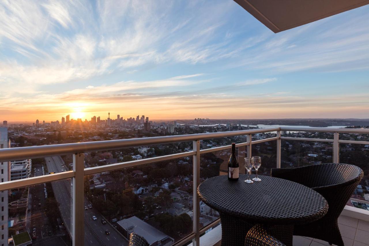 Meriton Suites Bondi Junction Sidney Dış mekan fotoğraf The image shows a scenic view from a balcony or terrace overlooking a cityscape during sunset. In the foreground, there is a small round table with two glasses and a bottle of wine, suggesting a romantic or relaxing setting. The sky is filled with vi