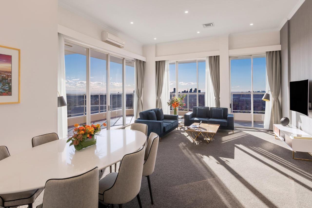 Meriton Suites Bondi Junction Sidney Dış mekan fotoğraf The photo shows a modern and stylish living space with large windows allowing natural light to fill the room. In the foreground, there is a round dining table with several chairs around it, adorned with a floral centerpiece. The seating area features
