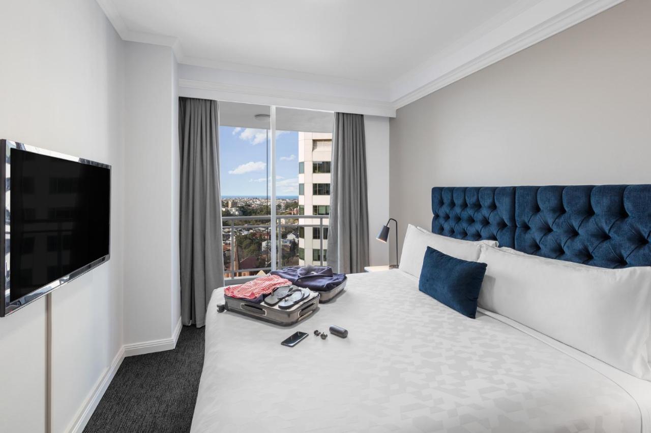Meriton Suites Bondi Junction Sidney Dış mekan fotoğraf The photo shows a modern hotel room featuring a neatly made bed with a blue tufted headboard and a white duvet. There's a small decorative pillow on the bed, and a piece of luggage is placed at the foot with clothes partially unpacked. A television i