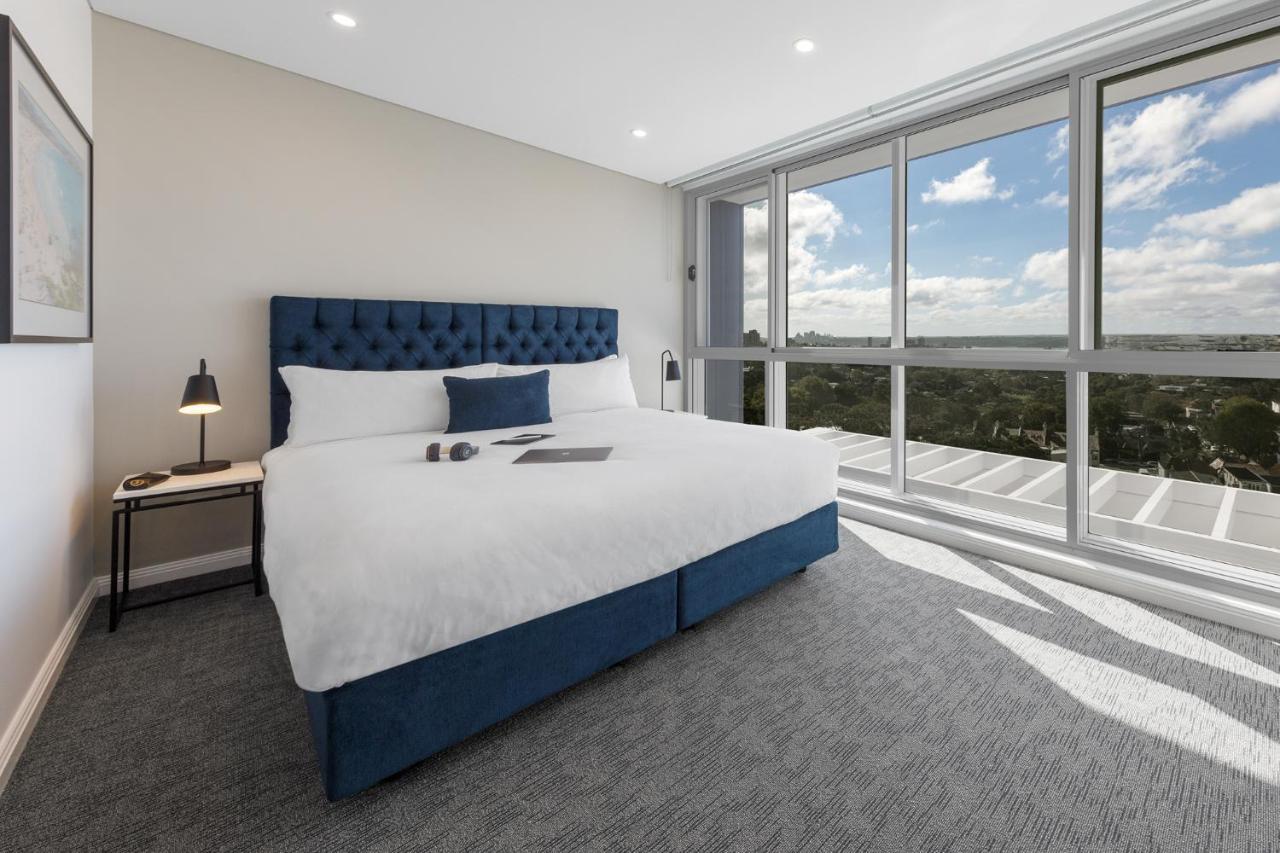 Meriton Suites Bondi Junction Sidney Dış mekan fotoğraf The photo shows a modern and stylish hotel room. It features a comfortable queen-size bed with a blue tufted headboard. The bed is neatly made with white linens and pillows. Next to the bed, there is a small bedside table with a desk lamp. 

On the f