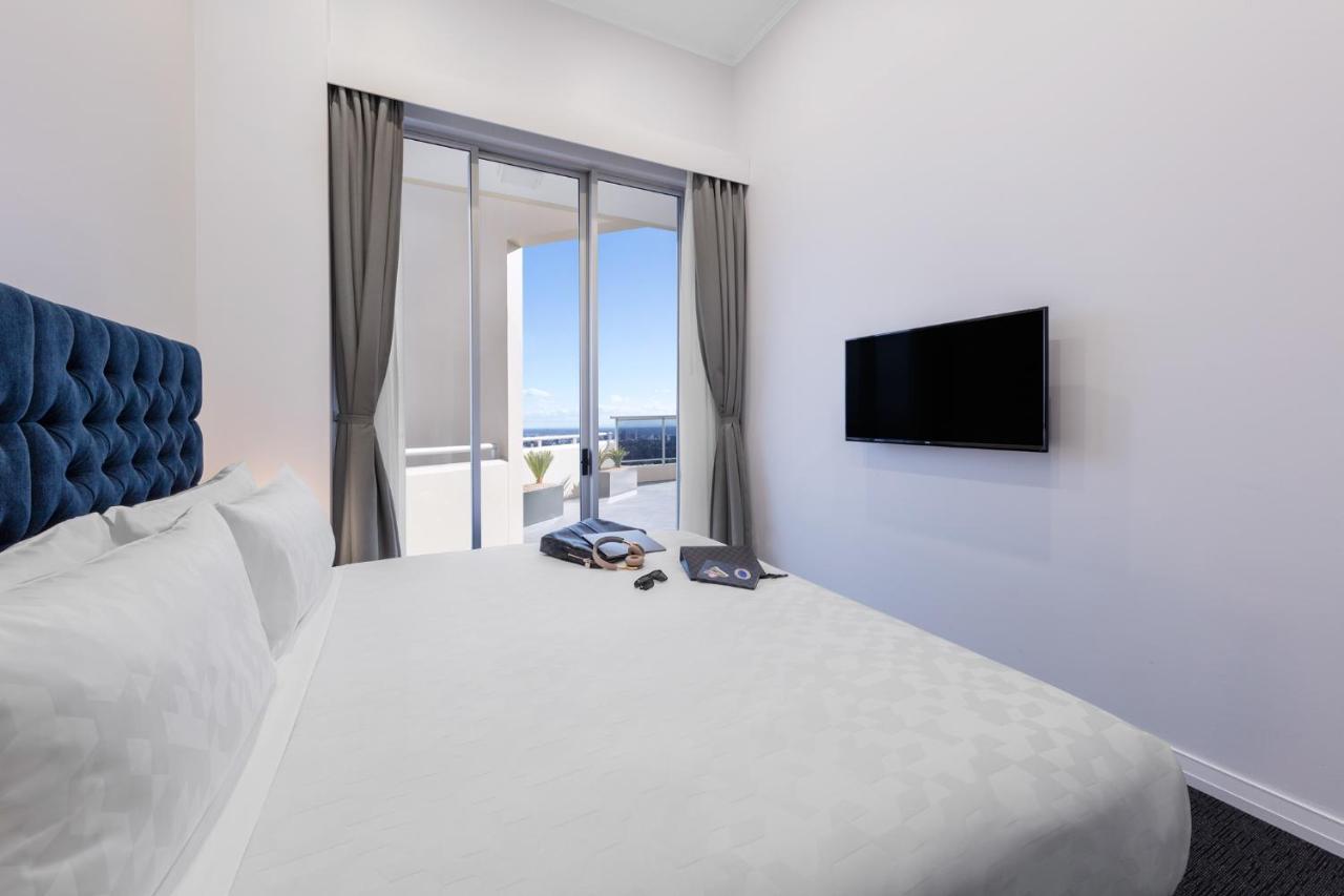 Meriton Suites Bondi Junction Sidney Dış mekan fotoğraf The photo shows a modern hotel room with a large, comfortable bed featuring a blue tufted headboard. There are light-colored walls, and the room is decorated simply. A flat-screen TV is mounted on the wall opposite the bed. The room has large glass d
