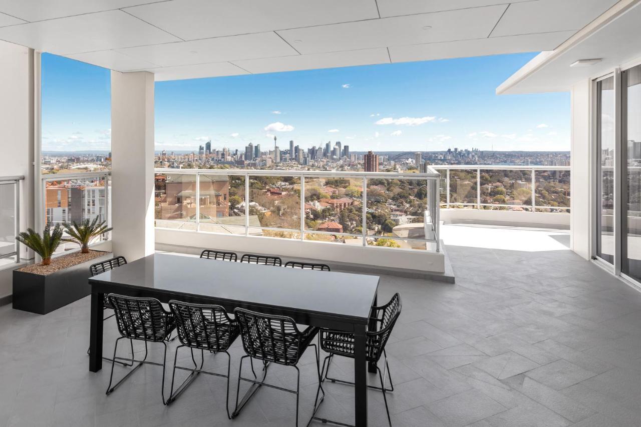 Meriton Suites Bondi Junction Sidney Dış mekan fotoğraf The photo shows a modern outdoor balcony or terrace space with a dining table and chairs. The table is black and has a minimalist design, surrounded by stylish chairs. In the background, there is a stunning view of a city skyline under a clear blue s