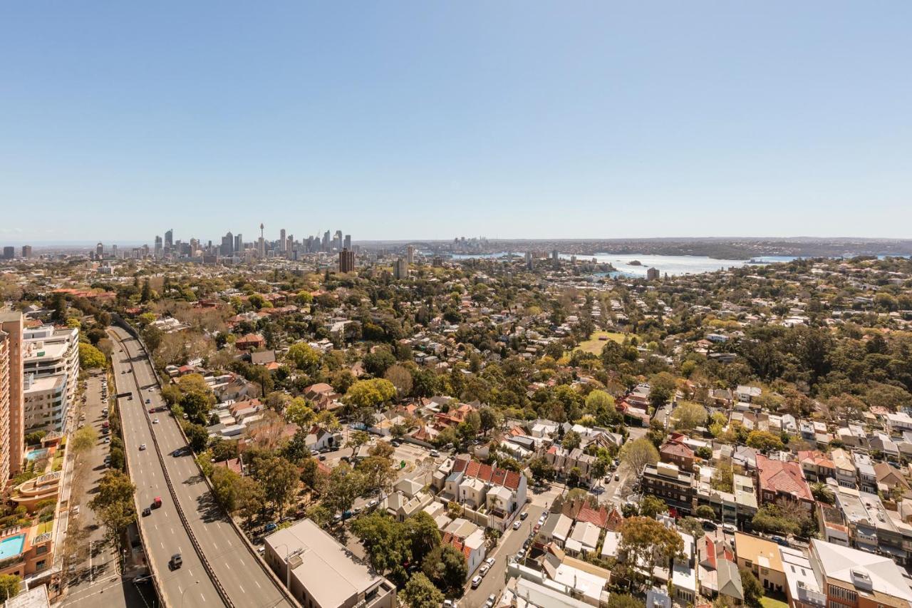 Meriton Suites Bondi Junction Sidney Dış mekan fotoğraf The photo captures a panoramic view of a vibrant urban landscape. In the foreground, there are residential areas with various houses and greenery, showcasing a suburban environment. In the background, the skyline of a city can be seen, featuring tall