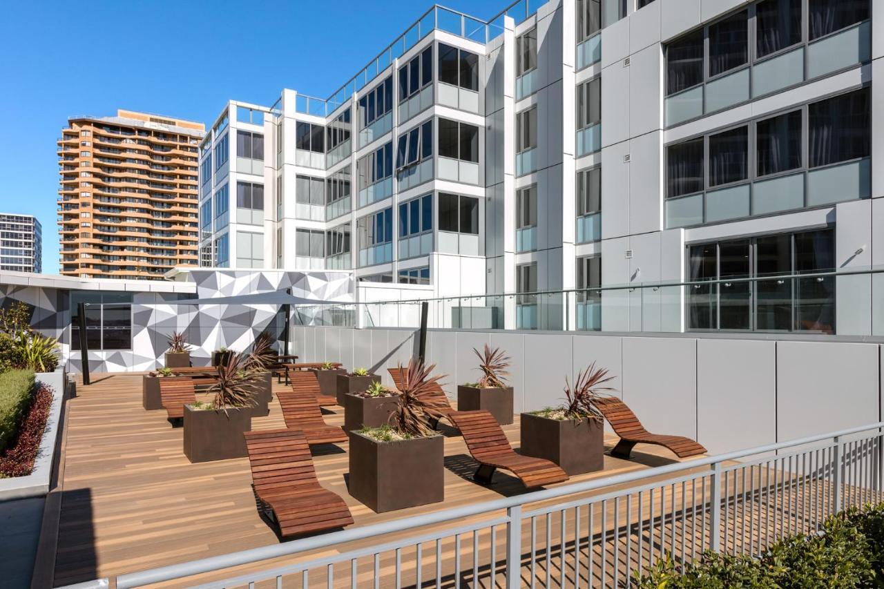 Meriton Suites Bondi Junction Sidney Dış mekan fotoğraf The photo shows a modern outdoor space featuring a wooden deck. There are several lounge chairs arranged around large planter boxes, which contain plants. In the background, there are contemporary residential buildings with large windows, and a geome
