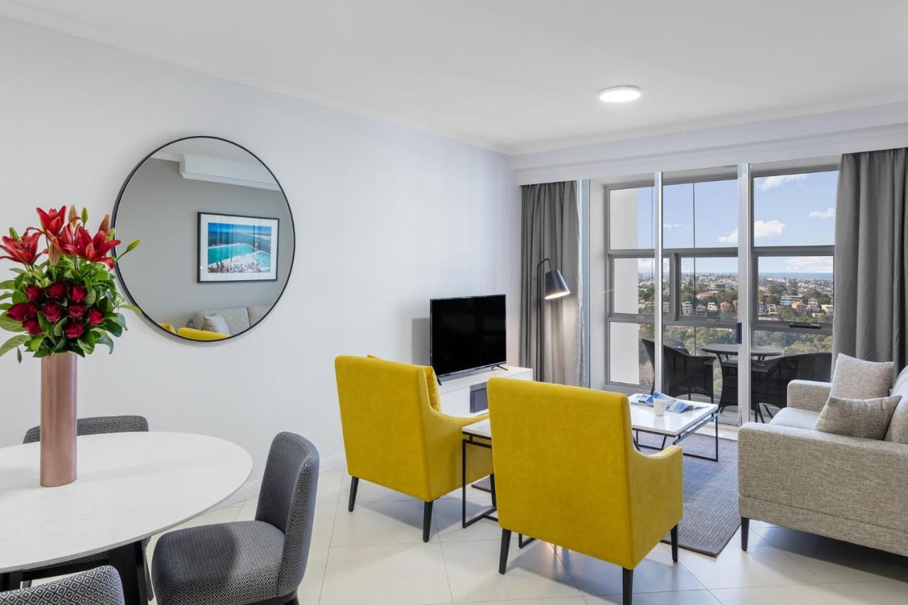 Meriton Suites Bondi Junction Sidney Dış mekan fotoğraf The photo shows a modern living space with a stylish design. There are two bright yellow chairs facing a small glass table, and a comfortable beige sofa to the right. A TV is positioned on the wall in front of the chairs. To the left, there's a dinin