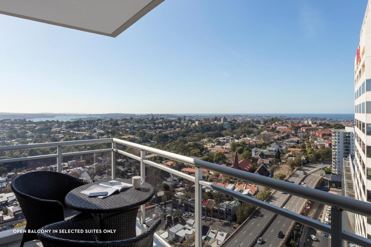 Meriton Suites Bondi Junction Sidney Dış mekan fotoğraf The photo shows a balcony with a view over a cityscape that includes buildings and greenery, extending towards the horizon where the sea is visible. The balcony features a small round table and two black chairs, suggesting a space for relaxation or d