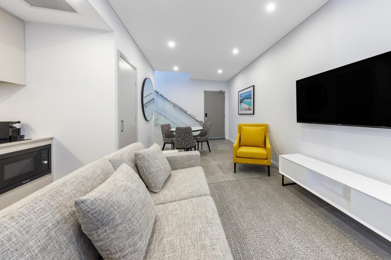 Meriton Suites Bondi Junction Sidney Dış mekan fotoğraf The photo shows a modern and stylish living space. It features a light-colored sofa with cushions, positioned to face a wall-mounted flat-screen TV. There's a yellow accent chair nearby that adds a pop of color to the room. The décor is minimalistic,