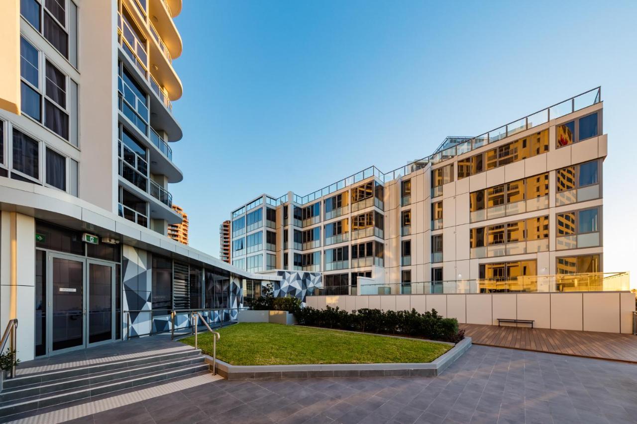 Meriton Suites Bondi Junction Sidney Dış mekan fotoğraf The photo showcases a modern residential area featuring contemporary buildings. In the foreground, there is a landscaped green space surrounded by well-designed architecture. The buildings have large glass windows, reflecting the light, and feature a