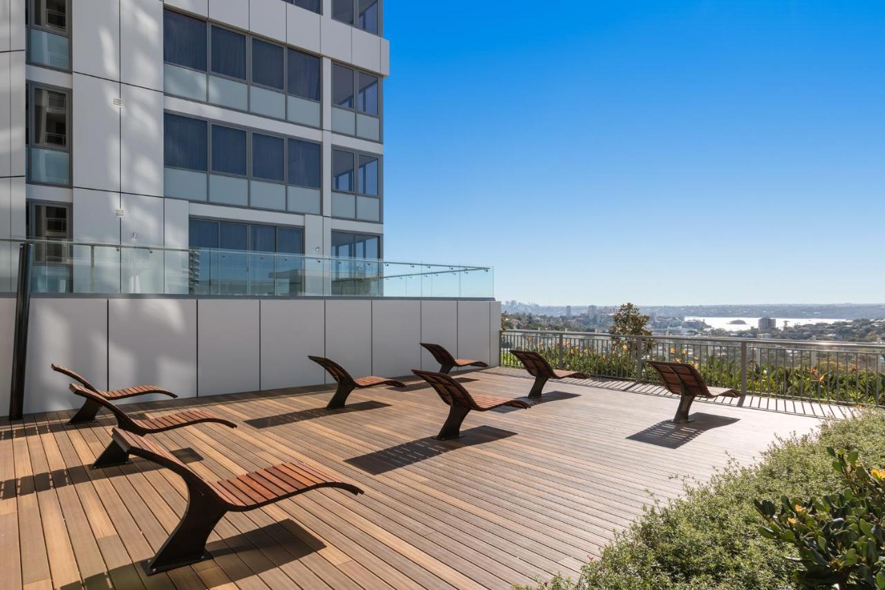 Meriton Suites Bondi Junction Sidney Dış mekan fotoğraf The photo shows a rooftop or outdoor area with a wooden deck. There are several modern lounge chairs arranged neatly, facing a clear sky and a panoramic view of the surrounding landscape. In the background, there is a tall building with large windows