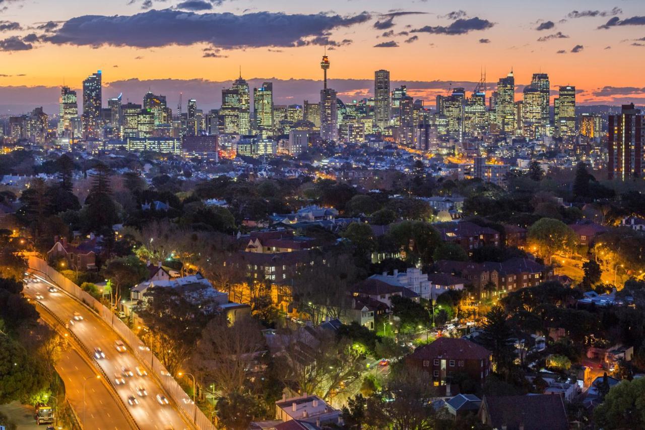 Meriton Suites Bondi Junction Sidney Dış mekan fotoğraf The photo captures a stunning city skyline at dusk, featuring towering skyscrapers adorned with lights against a colorful sunset backdrop. In the foreground, a green area with trees and houses is visible, while a road with cars can be seen winding th