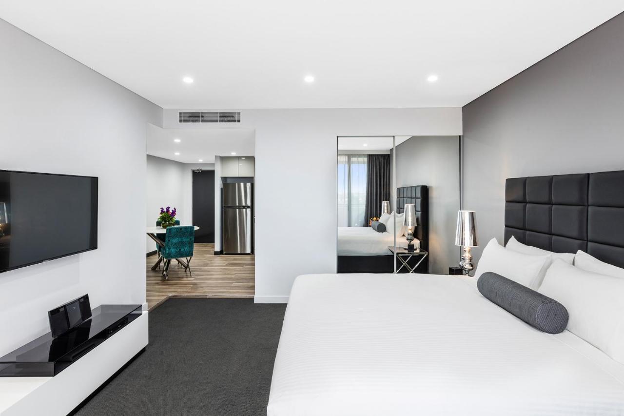 Meriton Suites Bondi Junction Sidney Dış mekan fotoğraf The photo shows a modern hotel room. It features a large bed with a white covering and gray accent pillows. To the left, there's a flat-screen TV mounted on the wall above a sleek white cabinet. The flooring is a light wood tone, and a large mirror i