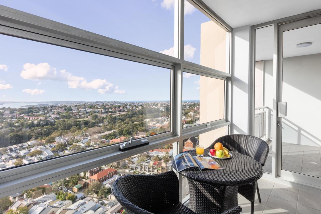 Meriton Suites Bondi Junction Sidney Dış mekan fotoğraf The photo shows a modern balcony with large glass windows that offer a panoramic view of a sprawling urban landscape. There is a small round table made of dark wicker, accompanied by two matching chairs. On the table, there is a bowl of fruit and a m