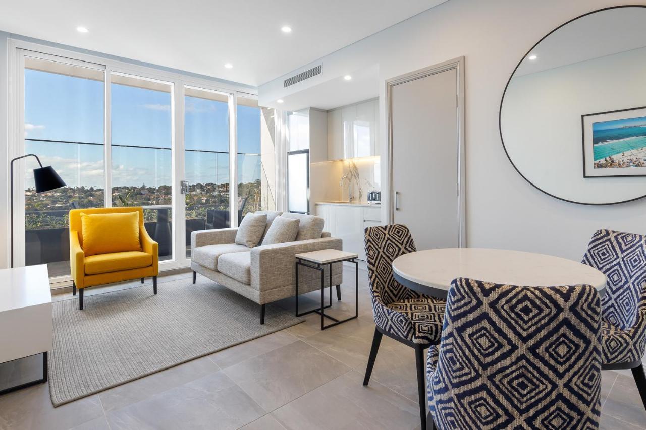 Meriton Suites Bondi Junction Sidney Dış mekan fotoğraf The photo shows a modern and stylish living area. It features a comfortable sofa in a neutral color, accompanied by a bright yellow armchair that adds a pop of color. There's a round dining table with patterned chairs nearby. Large windows allow plen