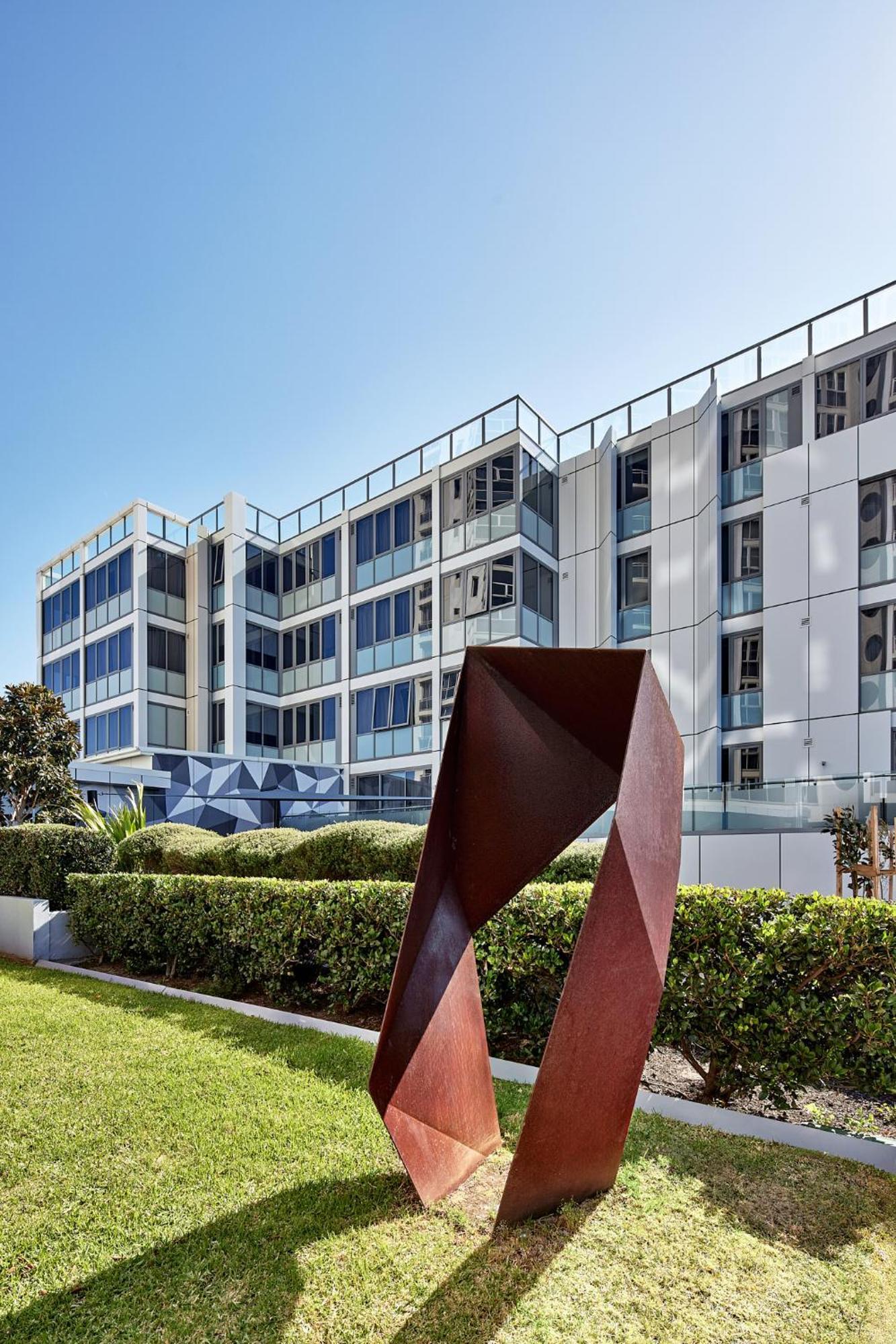 Meriton Suites Bondi Junction Sidney Dış mekan fotoğraf The photo shows a modern building with a sleek white and glass facade, featuring multiple floors and large windows. In the foreground, there is a sculptural piece made of metal, characterized by geometric shapes and a reddish-brown color. The surroun