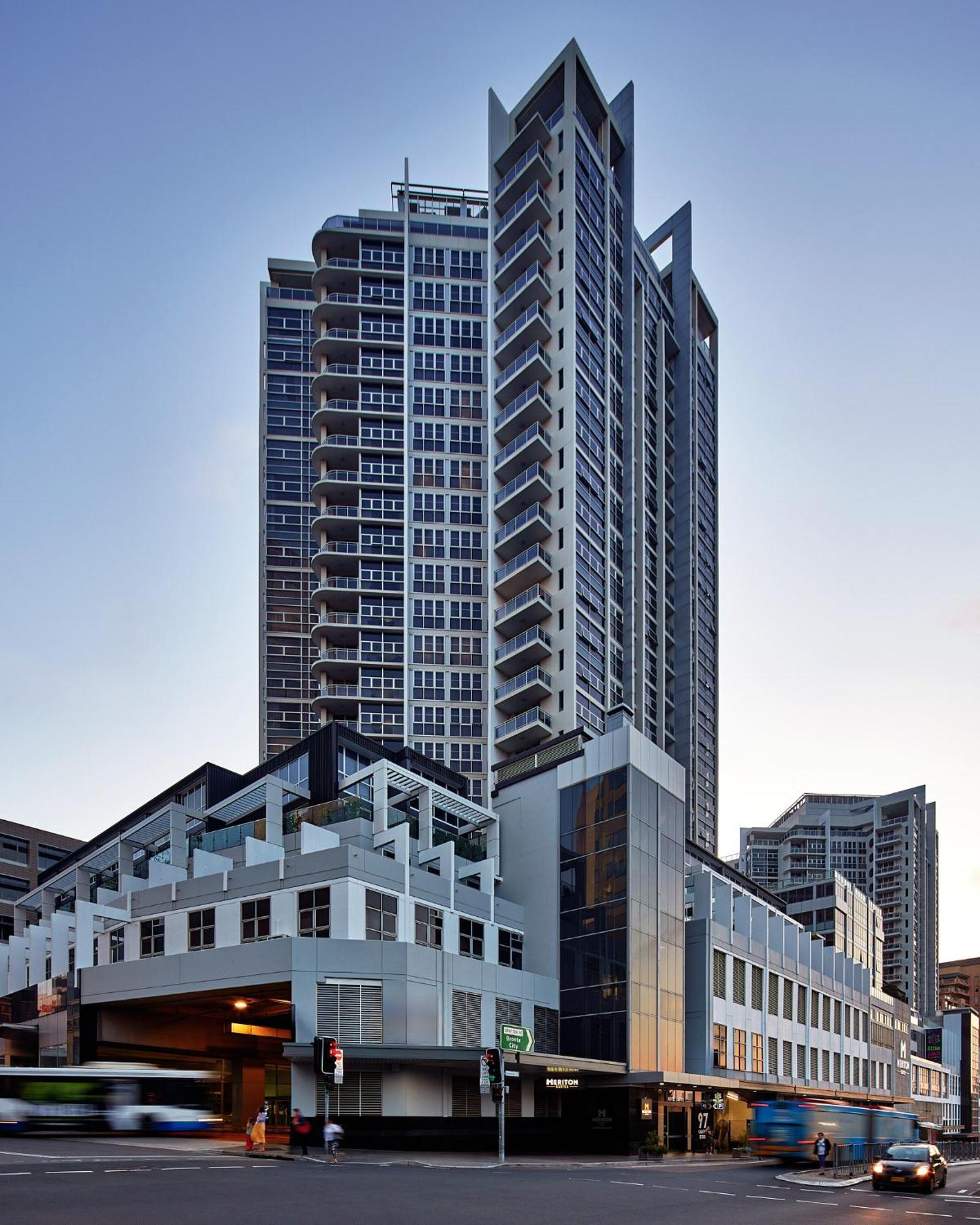 Meriton Suites Bondi Junction Sidney Dış mekan fotoğraf The image shows a modern high-rise building with a sleek design. The structure features a mix of glass and concrete elements, with multiple levels showcasing balconies. The building appears to be part of an urban environment, likely containing reside