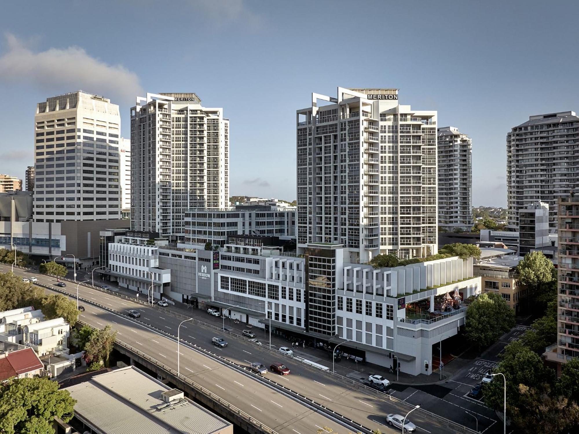 Meriton Suites Bondi Junction Sidney Dış mekan fotoğraf The photo shows a cityscape featuring several tall modern buildings, likely apartments or office complexes. In the foreground, there is a road lined with cars, and the architecture of the buildings varies, with some having sleek glass facades and oth
