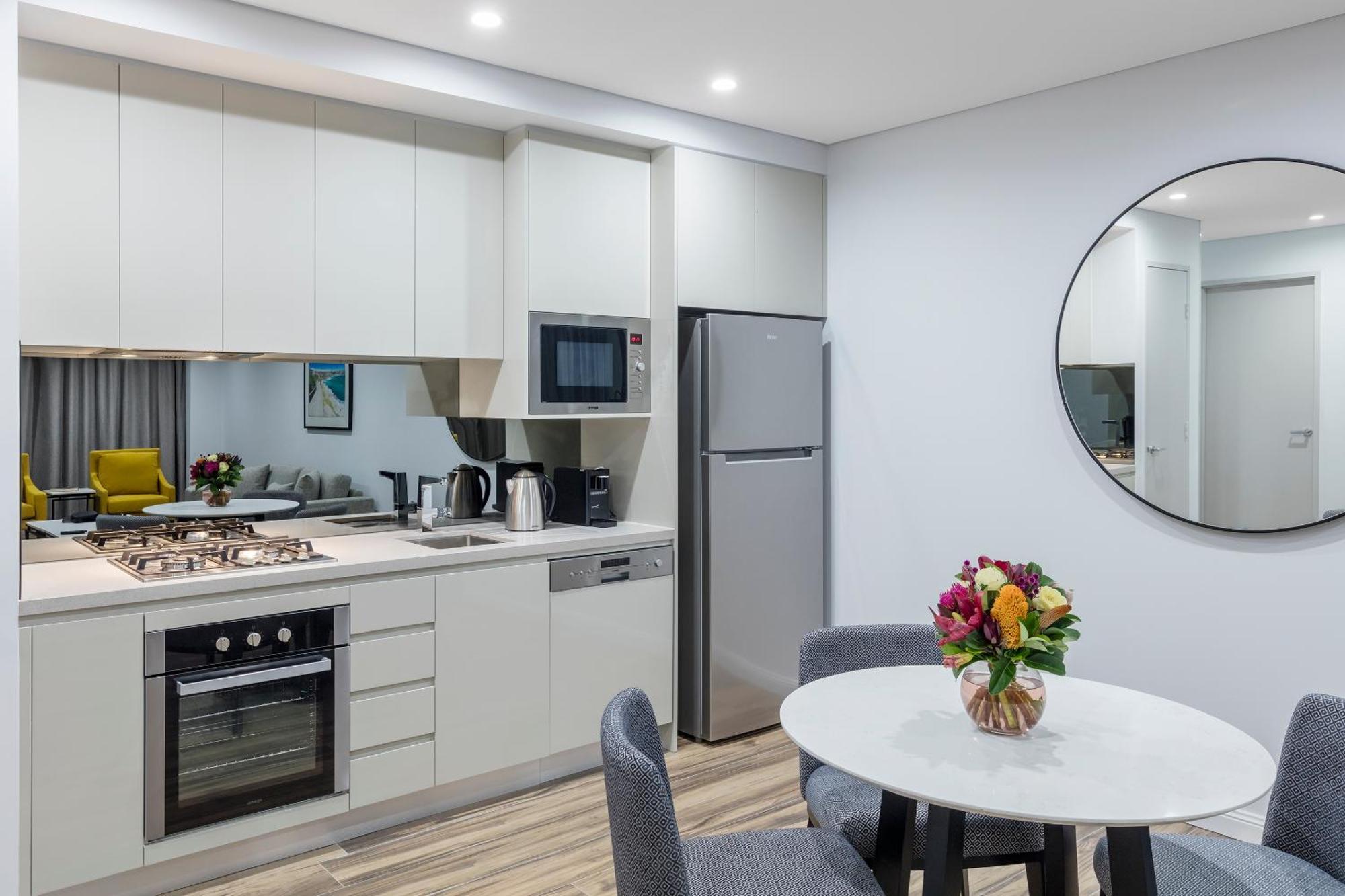Meriton Suites Bondi Junction Sidney Dış mekan fotoğraf The photo shows a modern kitchen and dining area. The kitchen features light-colored cabinetry with a built-in oven and stovetop, a microwave, and a silver refrigerator. There is a kettle and a coffee maker on the countertop. In the dining area, ther